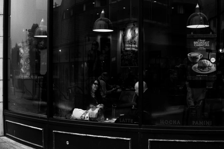 a black and white photo of a store front, a black and white photo, inspired by Brassaï, pexels contest winner, people inside eating meals, evening mood, drinking a coffee, light through glass