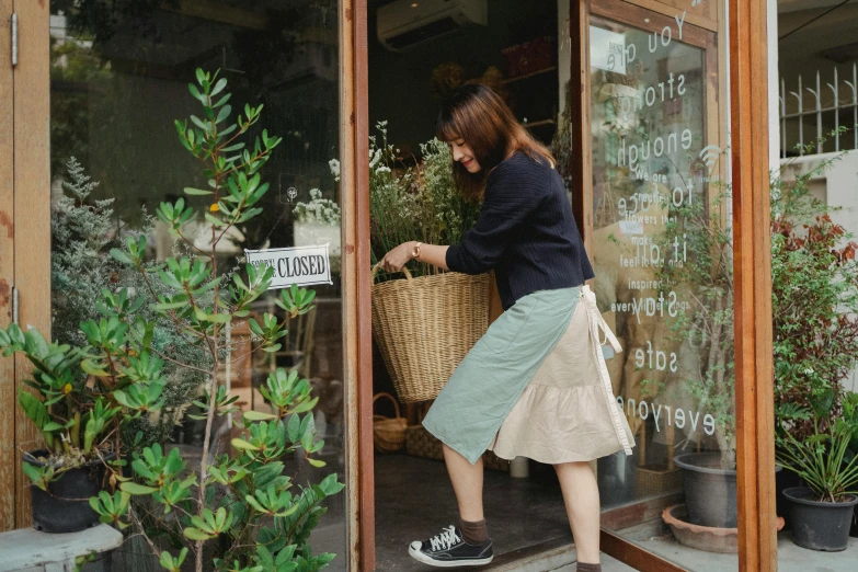 a woman standing at the door of a flower shop, inspired by Sawa Sekkyō, unsplash, white shirt and green skirt, with an easter basket, working clothes, grey