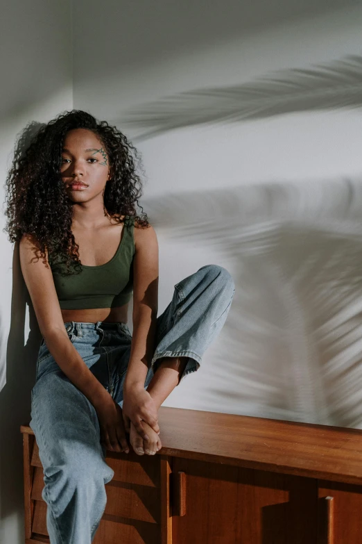 a woman sitting on top of a wooden bench, by Leo Michelson, trending on pexels, renaissance, black teenage girl, in white room, casual green clothing, curls on top