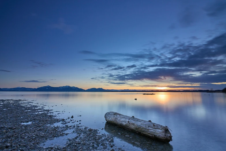 a log sitting on the shore of a lake, by Chris Rallis, unsplash contest winner, romanticism, blue sky at sunset, vancouver, calm sea, new zealand