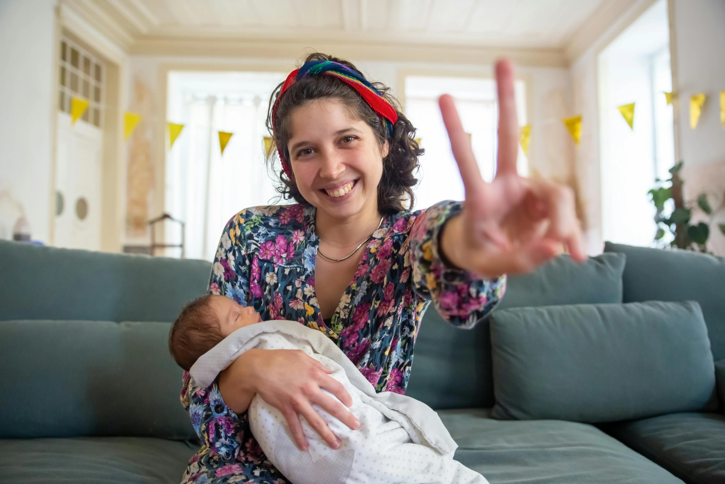 a woman sitting on a couch holding a baby, shutterstock contest winner, happening, peace sign, yael shelbia, close up portrait photo, waving and smiling