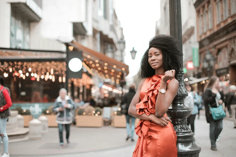 a woman standing next to a lamp post on a city street, by Chinwe Chukwuogo-Roy, pexels contest winner, happening, african american elegant girl, wearing festive clothing, orange hue, french girl