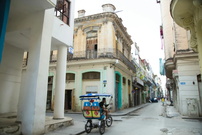 a man riding a bike down a street next to tall buildings, by Ceferí Olivé, preserved historical, cuban setting, a cozy, cart