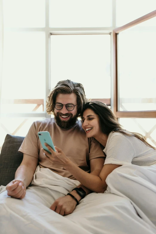 a man and woman sitting on a bed looking at a cell phone, happy vibes, on a couch, that's right, hug