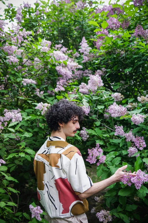 a man standing in front of a bush of purple flowers, an album cover, by Nina Hamnett, finn wolfhard, pale and coloured kimono, cottagecore flower garden, ignant