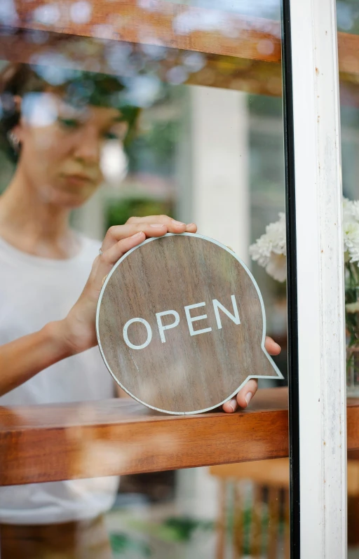 a woman holding an open sign in front of a window, pexels, square, shop front, brown, open plan