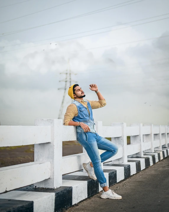 a man in overalls leaning against a fence, by Ismail Acar, trending on pexels, non binary model, assamese aesthetic, yellow cap, outfit : jeans and white vest
