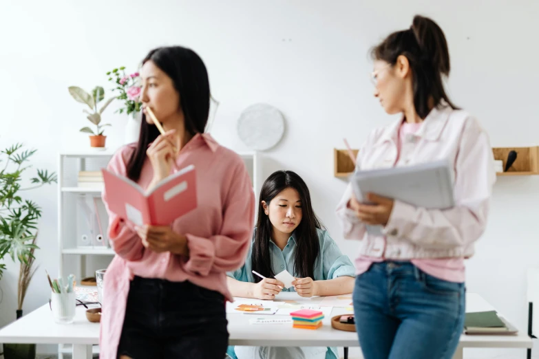 a group of women standing around a white table, trending on pexels, academic art, background image, holding notebook, young asian girl, three views