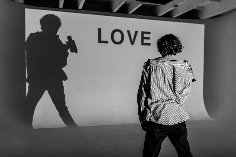 a man that is standing in front of a wall, a black and white photo, unsplash, graffiti, finn wolfhard, love in motion, low light museum, ed sheeran