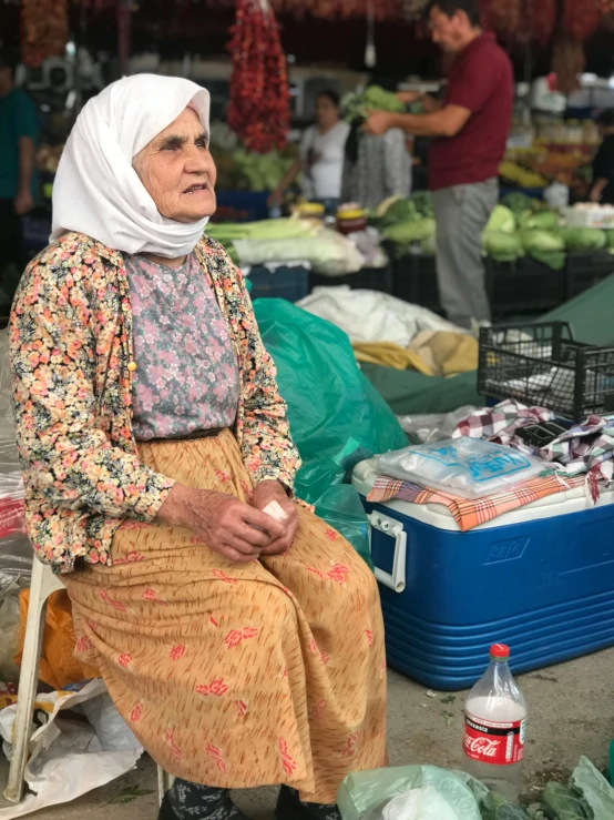 a woman sitting on a chair in a market, looks like ebru şahin, older woman, not smiling, square