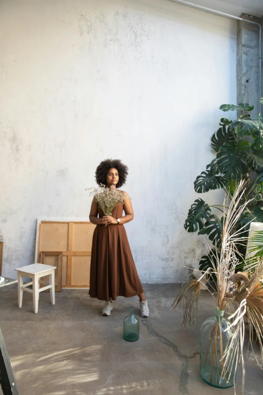 a woman in a brown dress standing in a room, inspired by Afewerk Tekle, trending on unsplash, with flowers and plants, curated collections, with afro, al fresco
