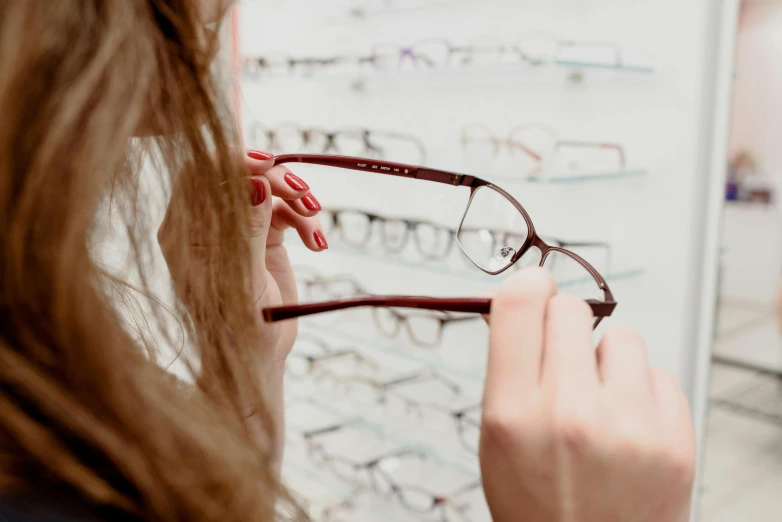 a woman holding a pair of glasses in front of a wall of glasses, trending on pexels, square rimmed glasses, inspect in inventory image, close-up shot from behind, with glasses on