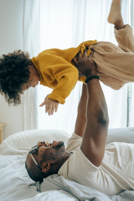 a man laying on top of a bed holding a child, inspired by The Family Circus, pexels contest winner, african american, soft shapes, basil flying, kids playing