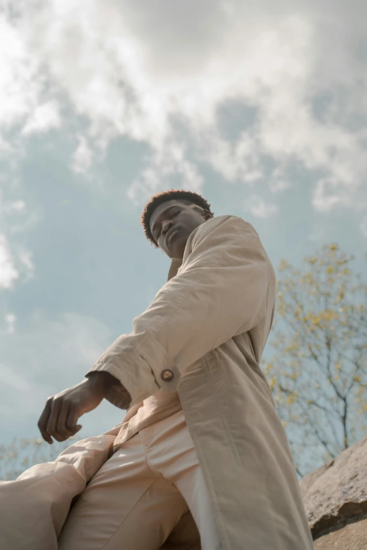 a man in a trench coat sitting on a rock, an album cover, trending on unsplash, happening, low angle facing sky, brown skinned, ( ( theatrical ) ), wearing a linen shirt