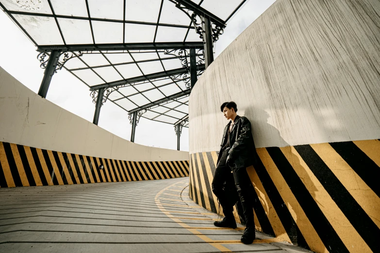 a man in a black jacket leaning against a wall, an album cover, inspired by Zhang Han, unsplash, visual art, overpass, pentagon, sky bridge, wide angle full body