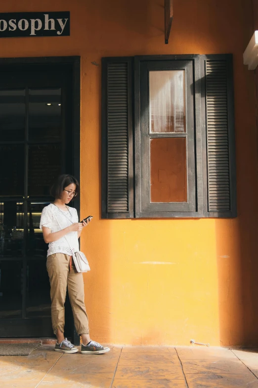 a woman standing outside of a building looking at her phone, inspired by Cheng Jiasui, pexels contest winner, pale orange colors, malaysian, shutters, soft shadow