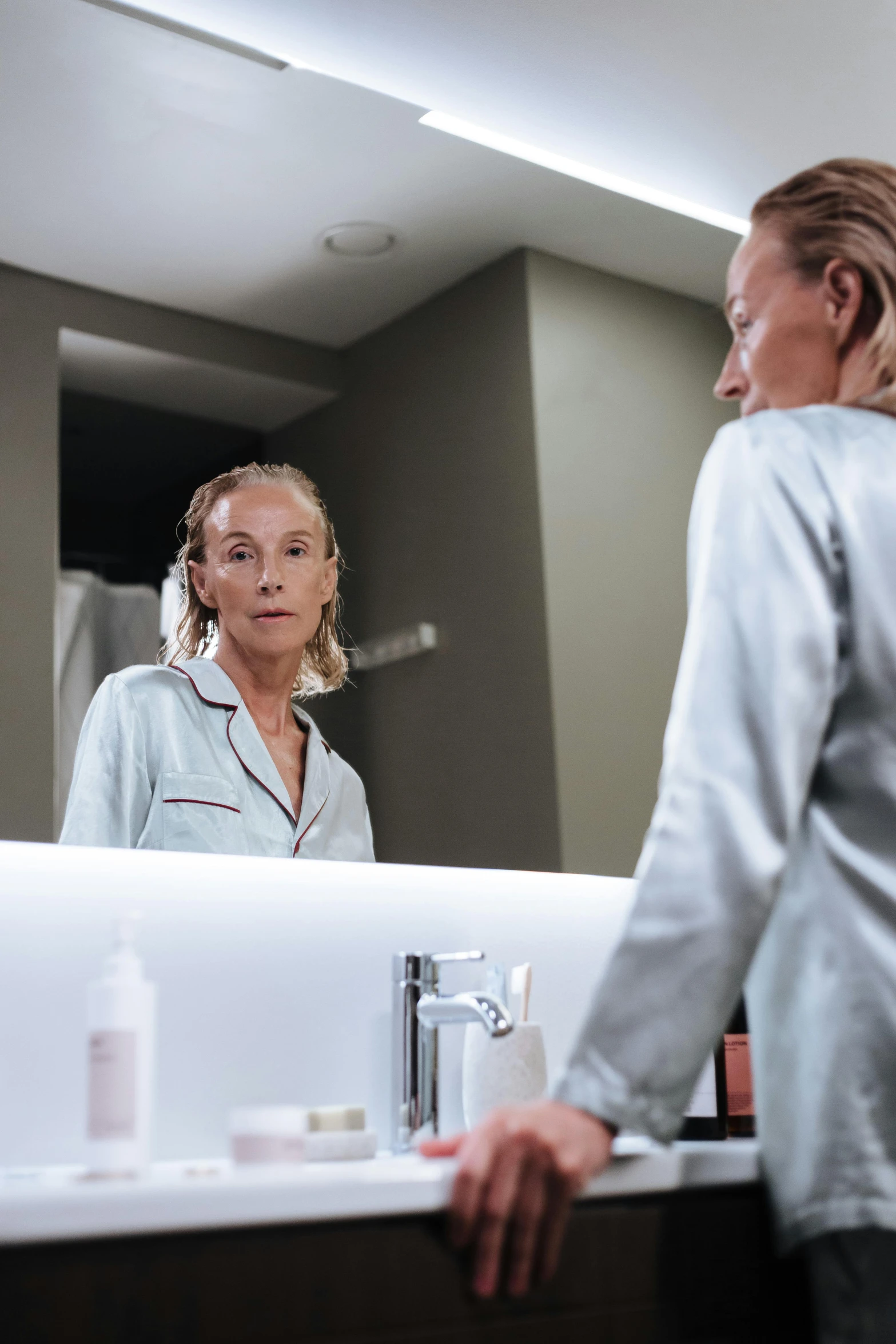 a woman standing in front of a bathroom mirror, happening, wearing a grey robe, 5 5 yo, facial precision, a blond