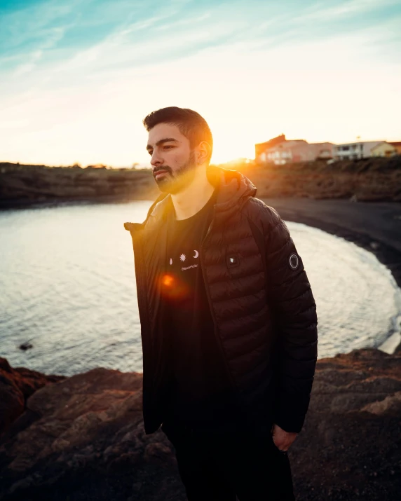 a man standing in front of a body of water, an album cover, pexels contest winner, happening, model wears a puffer jacket, sunset lighting 8k, irish youtuber, profile image