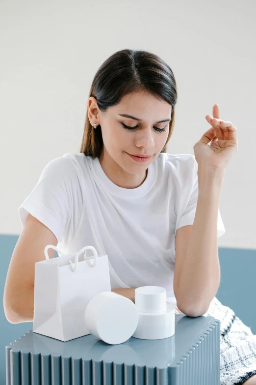 a woman sitting at a table with shopping bags, a marble sculpture, trending on pexels, minimalism, dressed in a white t-shirt, skincare, holding flask in hand, wearing a cute top