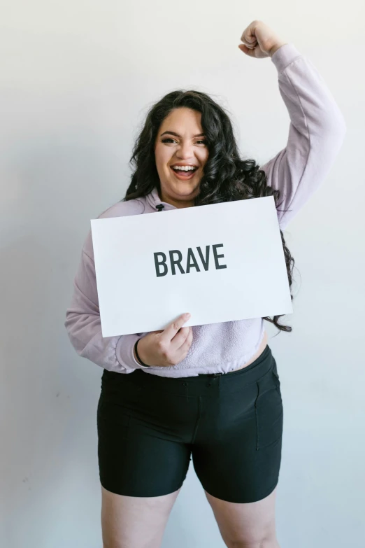 a woman holding a sign that says brave, by Byron Galvez, trending on unsplash, plus size, hero pose, on a pale background, excited