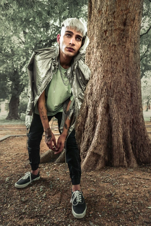 a man standing next to a tree in a park, an album cover, inspired by Byron Galvez, trending on pexels, hyperrealism, lil peep, green clothing, attractive androgynous humanoid, model is wearing techtical vest