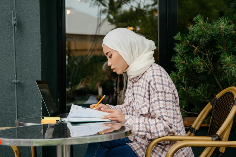 a woman sitting at a table writing on a piece of paper, a cartoon, inspired by Maryam Hashemi, pexels contest winner, hurufiyya, white hijab, at college, casual pose, holding notebook
