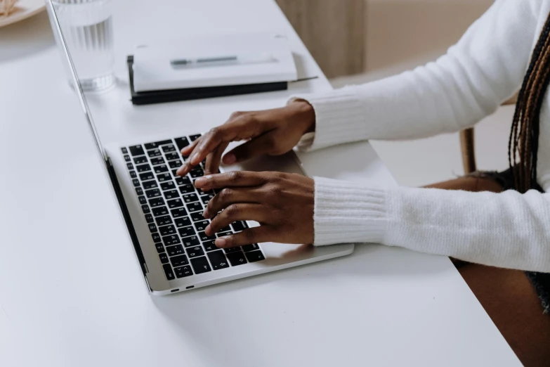 a woman sitting at a desk typing on a laptop, by Carey Morris, trending on pexels, wearing a white sweater, sitting with wrists together, riyahd cassiem, elegantly dressed