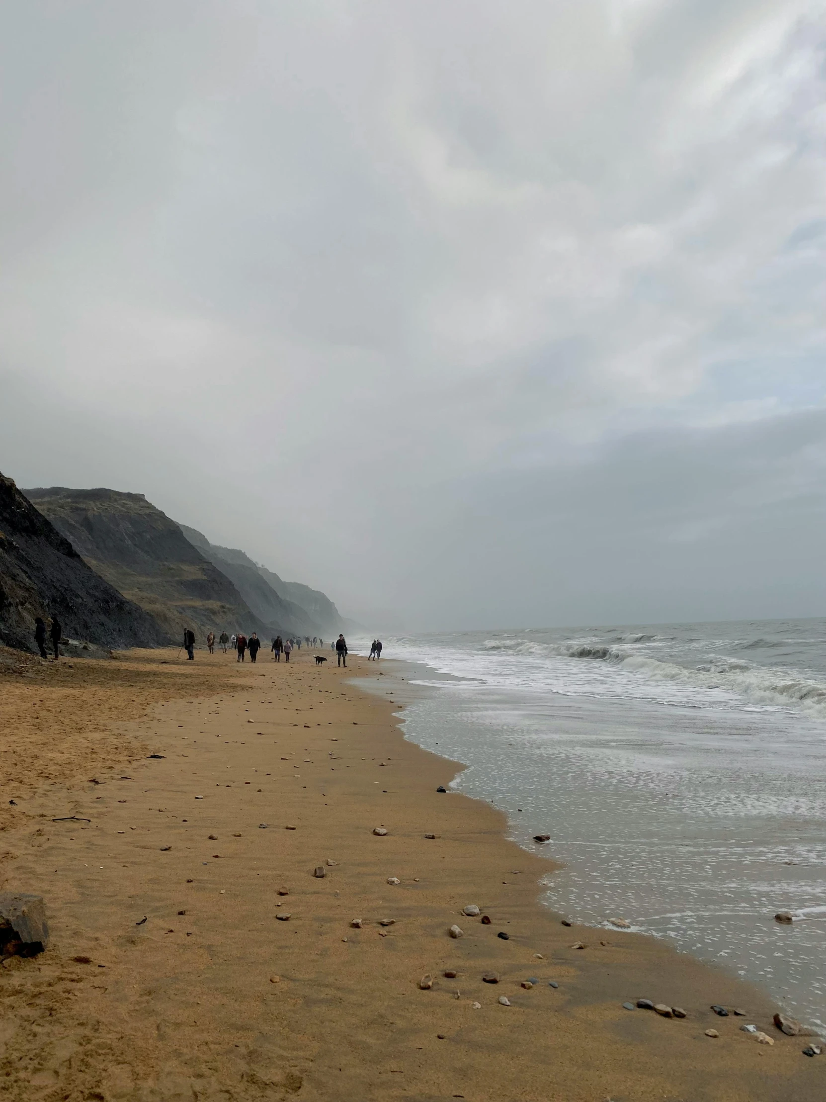 a group of people walking along a sandy beach, sea storm and big waves cliffs, grey skies, chalk cliffs above, about to step on you