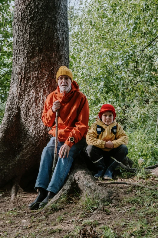 a couple of people sitting next to a tree, grandfatherly, wearing adventure gear, with a kid, wearing red and yellow clothes
