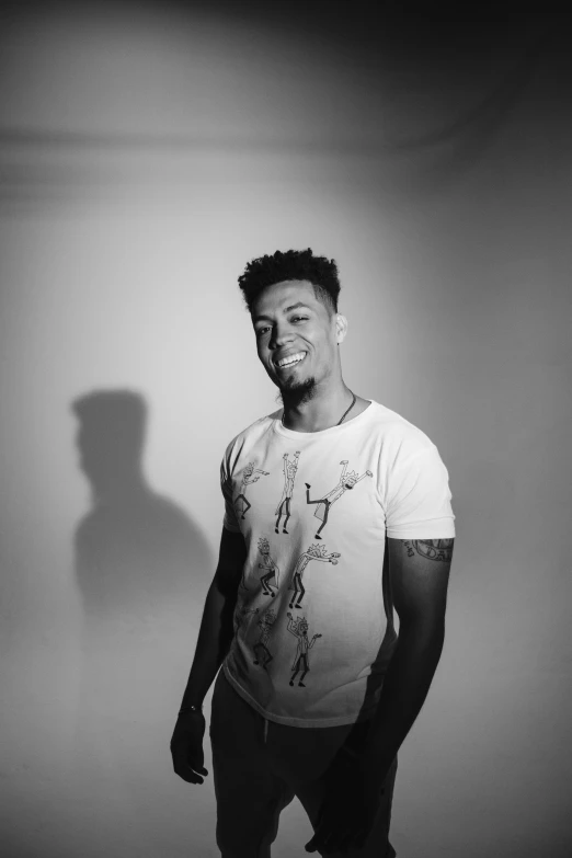 a man standing in front of a white wall, a black and white photo, inspired by Frank Mason, 2 1 savage, his smile threw shadows, dressed in a white t shirt, backlight studio lighting