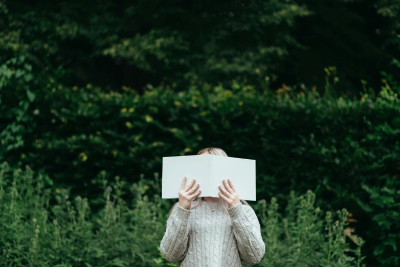 a woman covering her face with a book, a photo, by Carey Morris, pexels contest winner, wearing a white sweater, outdoor photo, minimalist photo, background image
