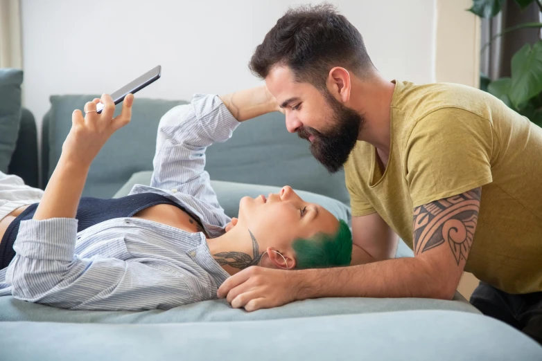 a man and a woman laying on a couch looking at a cell phone, a tattoo, trending on pexels, short green hair, couple on bed, raphael lecoste, tattoo artist