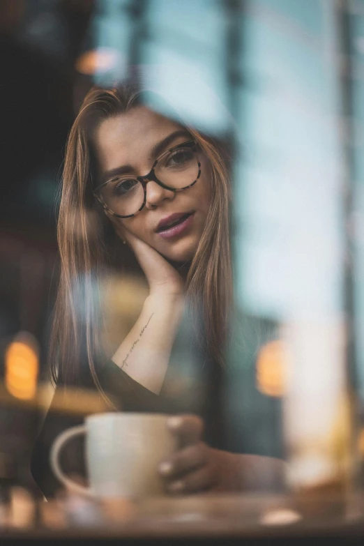 a woman sitting at a table with a cup of coffee, a picture, by Adam Marczyński, trending on pexels, renaissance, girl wearing round glasses, depressed girl portrait, square rimmed glasses, reflecting
