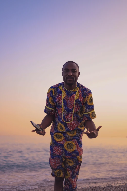 a man standing on top of a beach next to the ocean, an album cover, inspired by Ras Akyem, pexels contest winner, happening, wearing an african dress, pose(arms up + happy), still from a music video, african jesse pinkman portrait