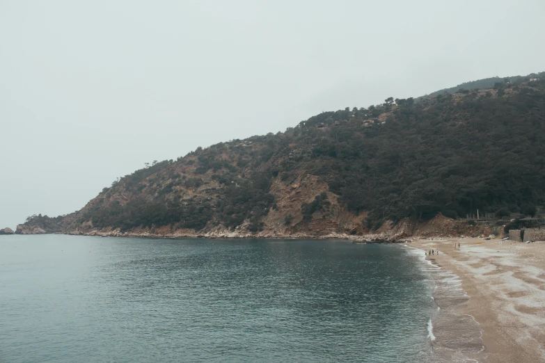 a sandy beach with a hill in the background, pexels contest winner, under a gray foggy sky, mediterranean beach background, view of forest, looking down a cliff