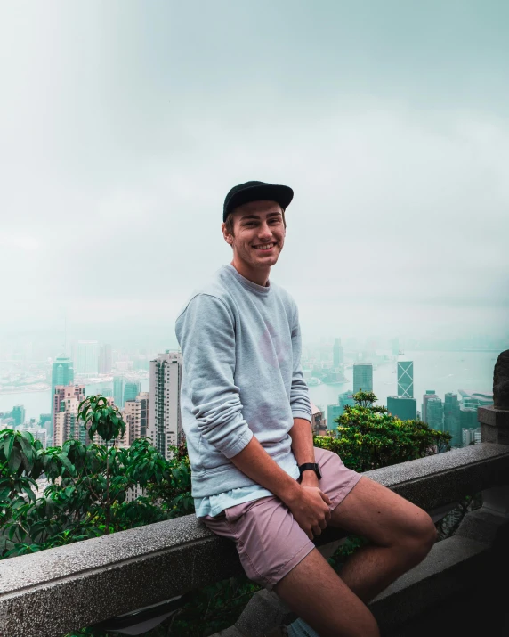 a man sitting on a ledge with a city in the background, happily smiling at the camera, in hong kong, lush surroundings, tinder profile