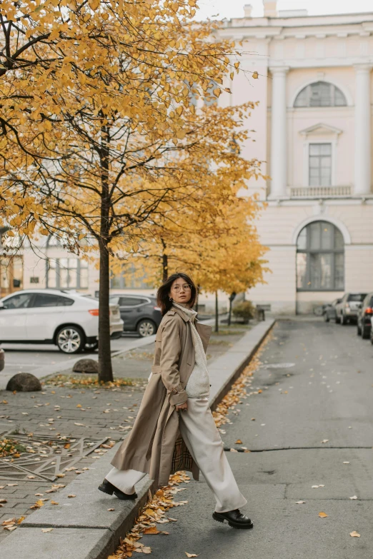 a woman walking down a sidewalk in front of a building, by Christen Dalsgaard, trending on unsplash, visual art, autumnal empress, long coat, finland, next to a tree