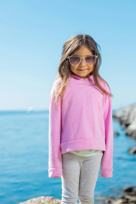 a little girl standing on a rock by the water, wearing a pastel pink hoodie, wearing shades, zoomed in, full product shot
