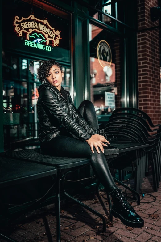 a woman sitting on a bench in front of a building, wearing a leather jacket, ashteroth, taverns nighttime lifestyle, leather body suit