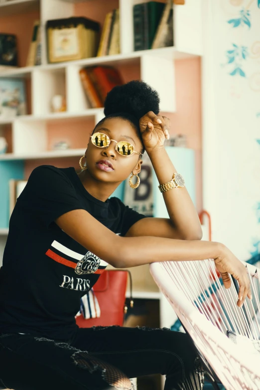 a woman sitting on a chair in a room, trending on pexels, afrofuturism, round sunglasses, graphic tees, gold accessories, willow smith young