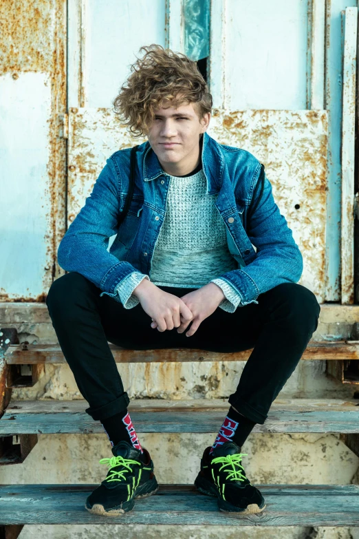 a young man sitting on the steps of a building, an album cover, by John Luke, curly blond, profile image, striking a pose, sitting on a stool