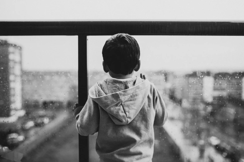 a black and white photo of a child looking out a window, pexels, on a balcony, wearing jeans and a black hoodie, cloudy day, 2019 trending photo