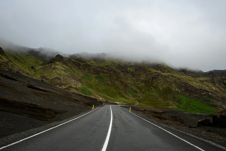 an empty road with a mountain in the background, by Hallsteinn Sigurðsson, pexels contest winner, hurufiyya, under a gray foggy sky, some of the blacktop is showing, green eays, craggy