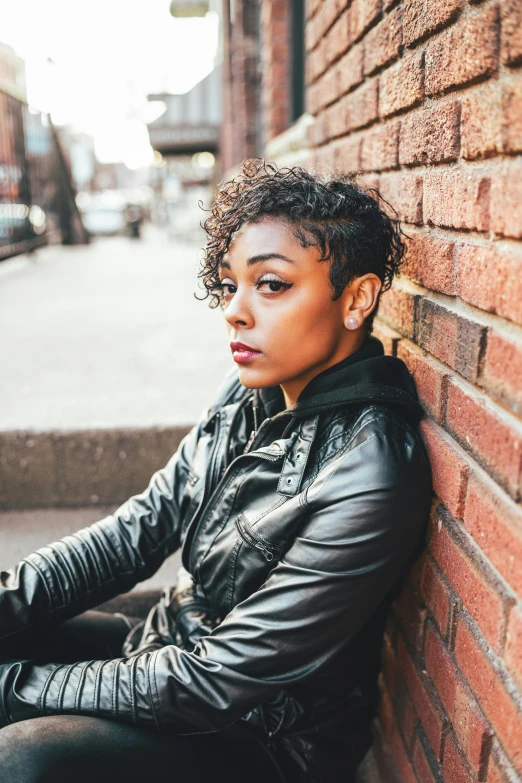 a woman sitting against a brick wall, by Jason Felix, trending on pexels, short black curly hair, wearing leather jacket, androgynous person, riyahd cassiem