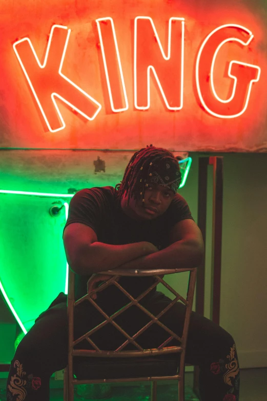 a man sitting on a chair in front of a neon sign, an album cover, trending on pexels, wearing kings crown, black teenage boy, thicc, ( ( theatrical ) )