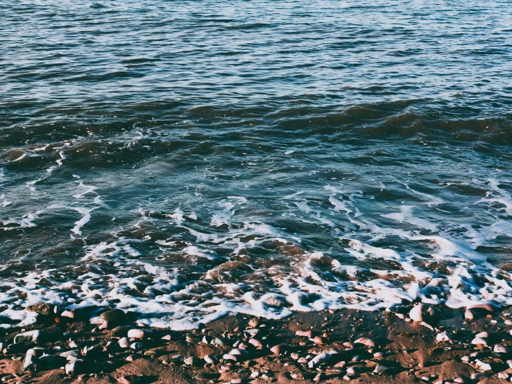 a person riding a surfboard on top of a sandy beach, an album cover, unsplash, water ripples, black sea, 1990's photo, rocky shore