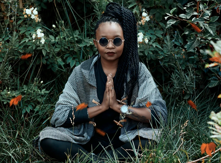 a woman sitting in the grass with her hands together, pexels contest winner, afrofuturism, wearing black rimmed glasses, praying posture, black lotus, african woman