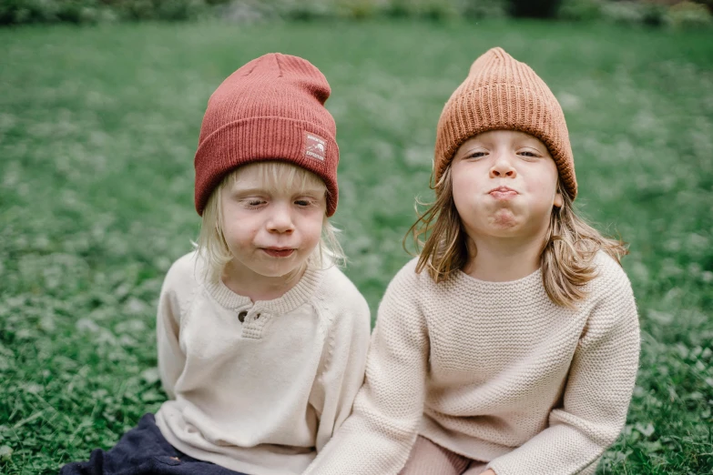 a couple of kids sitting on top of a lush green field, by Nina Hamnett, pexels contest winner, beanie hat, squinting, warm cosy colors, maroon