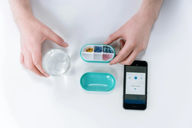 a person holding a container of food next to a cell phone, inspired by Damien Hirst, turquoise, product design, pills, digital medical equipment