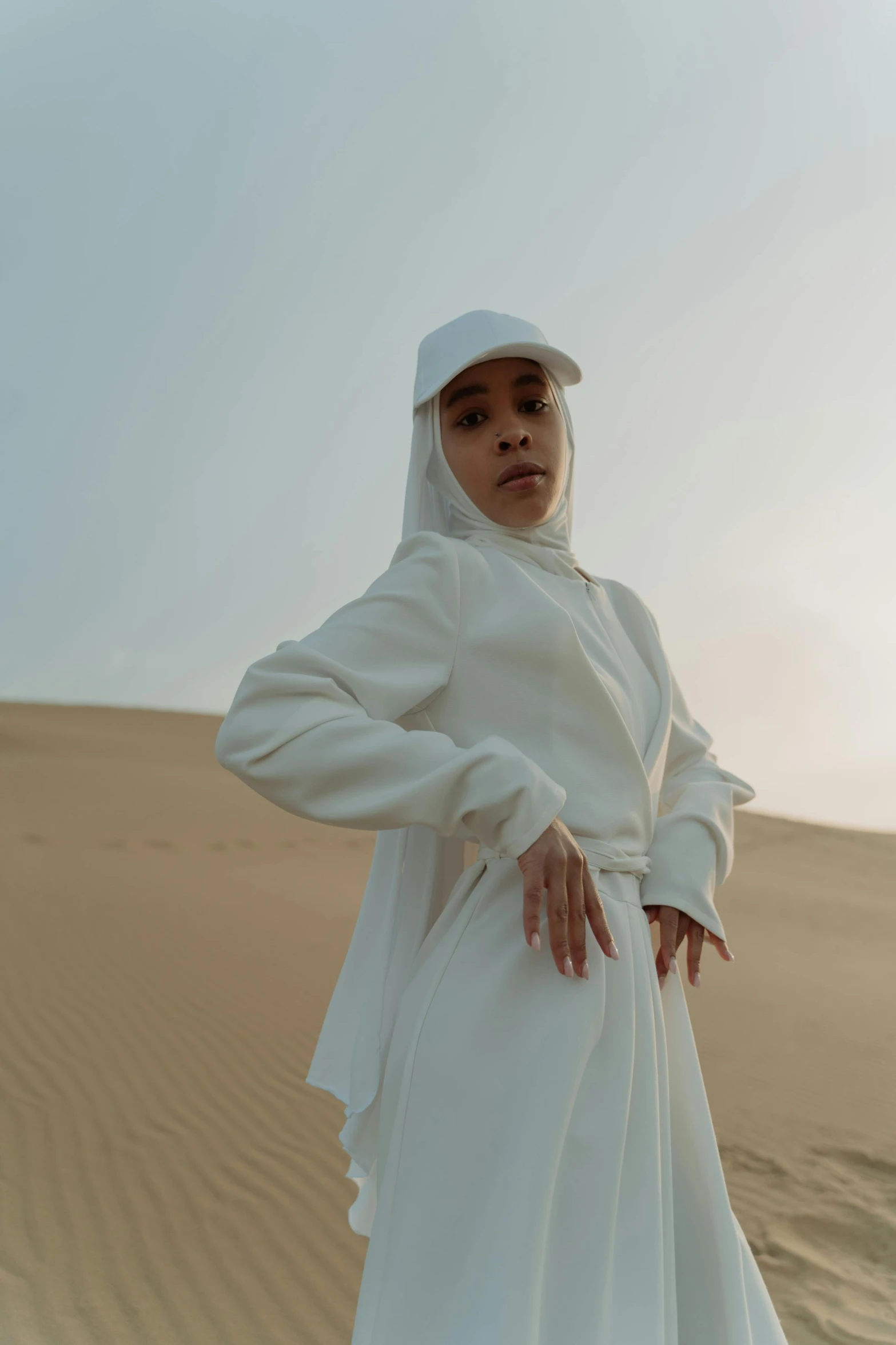 a woman in a white dress standing in the desert, an album cover, inspired by Amir Zand, trending on pexels, hurufiyya, wearing a hoodie and sweatpants, ferred - themed robes and hat, portrait mode photo, taken in the late 2010s